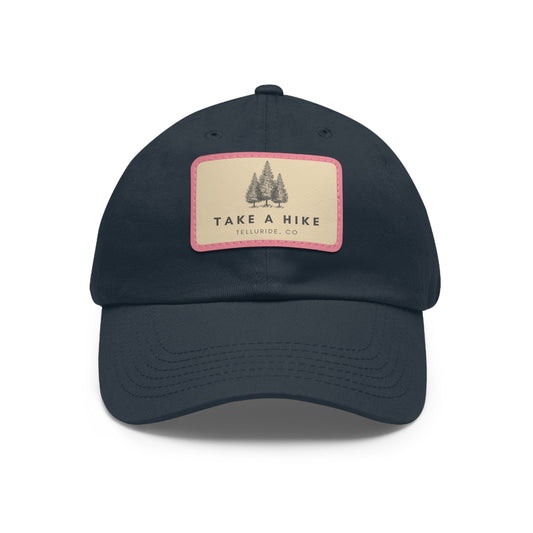 Navy dad hat with a brown leather patch with pink outline featuring black lettering saying "Take A Hike" and Telluride, CO. The hat is perfect for keeping the sun out of your face while you're hiking or skiing. The phrase "Take A Hike" is a popular saying among hikers and skiers, and the words "Telluride, CO." represent the beauty of the Telluride Ski Resort. This hat is a great way to show your love of the outdoors and your love of Telluride, Colorado.