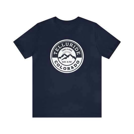 Unisex short sleeve t-shirt with a circle navy and white logo featuring white lettering saying "Telluride, Colorado", "Take A Hike", and "Elev. 8,750". The t-shirt is made of 100% cotton and is perfect for a day of hiking or skiing. The phrase "Take A Hike" is a popular saying among hikers and skiers, and the words "Telluride, Colorado" represent the beauty of the Telluride Ski Resort. This t-shirt is a great way to show your love of the outdoors and your love of Telluride.