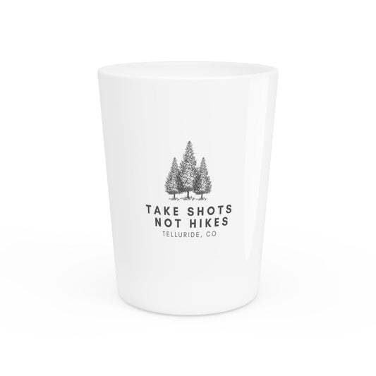 White ceramic shot glass with black lettering saying "Take Shots Not Hikes" and Telluride, CO. The shot glass is perfect for enjoying a shot of your favorite spirit at the Telluride Ski Resort or anywhere else you enjoy spending time in the great outdoors. The phrase "Take Shots Not Hikes" is a humorous way to express your love of skiing and snowboarding, and the words "Telluride, CO" represent the beauty of the Telluride Ski Resort. 