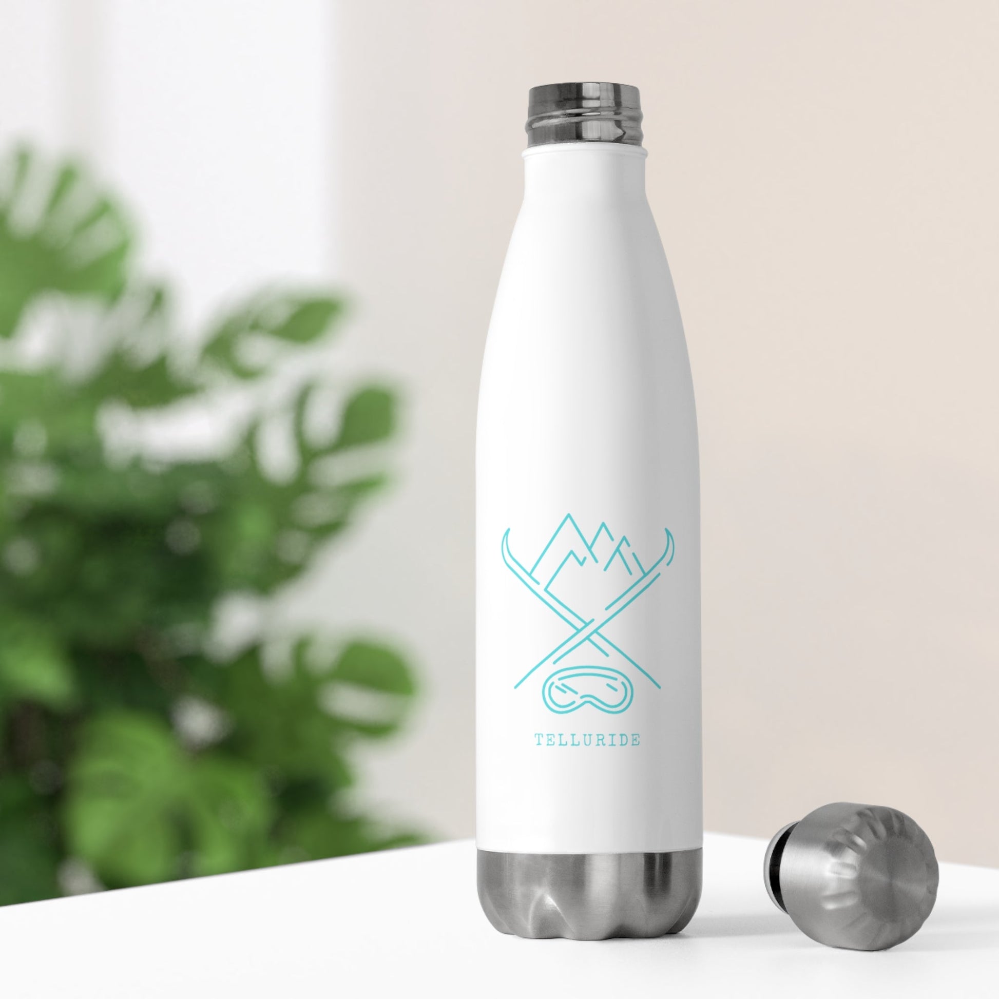 White stainless steel insulated 20oz bottle with a teal image of crossed skis, mountains, and ski goggles. The word "Telluride" is printed in teal on the front of the bottle. The bottle is a stylish and functional way to stay hydrated and show your love of skiing and Telluride. The white bottle is simple and elegant, while the teal image and lettering add a touch of color. The bottle is a great way to enjoy a cold drink on a hot day or to take with you on your next ski trip to the Telluride Ski Resort.